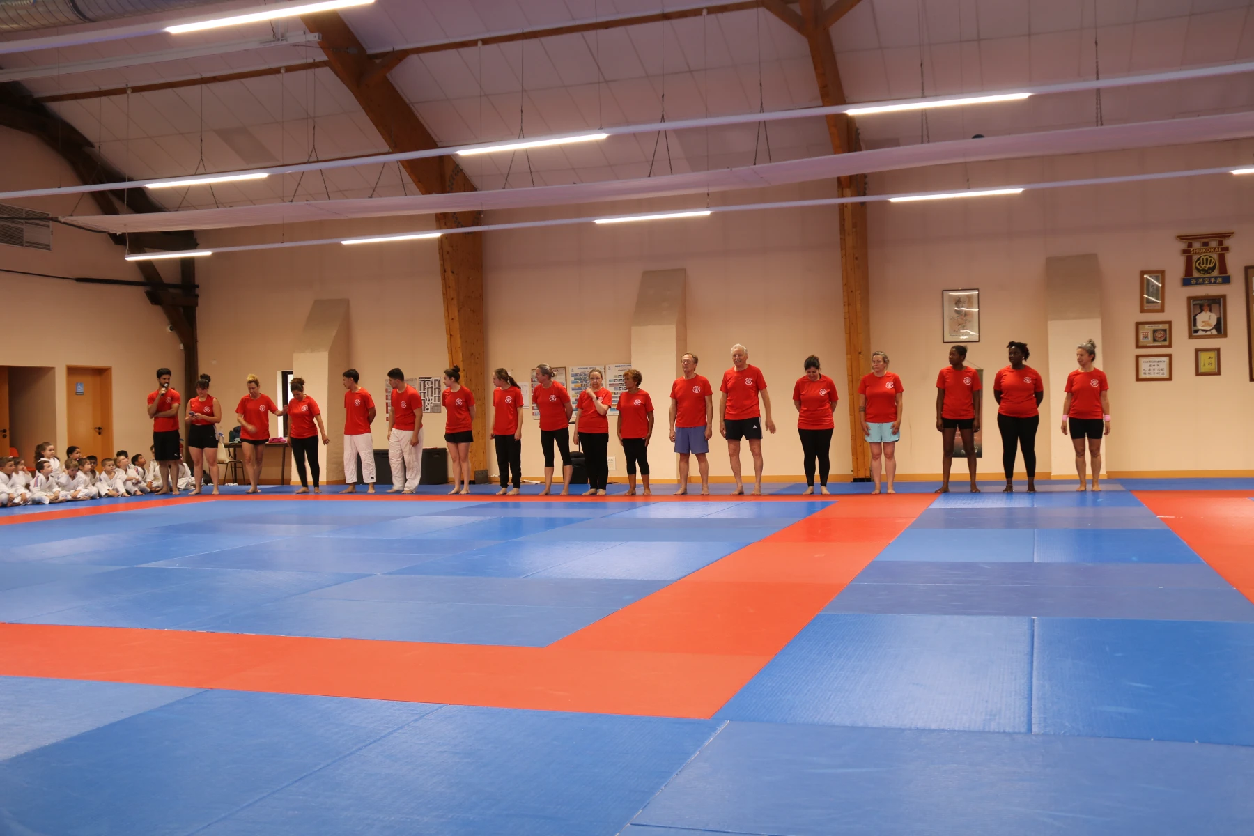 Les animateurs du club en t-shirt rouge, alignés sur le tatami, s'apprêtent à lancer une démonstration devant les enfants assis.