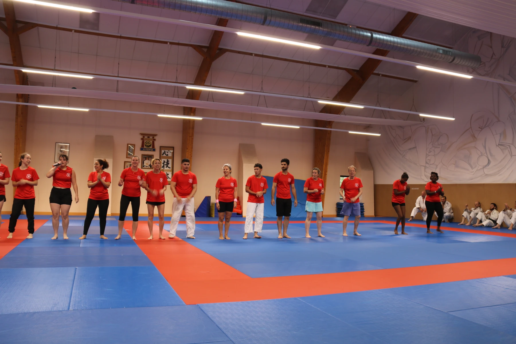 Groupe d'animateurs en t-shirt rouge alignés et souriants, l'un au micro, lors d'une animation festive au dojo.