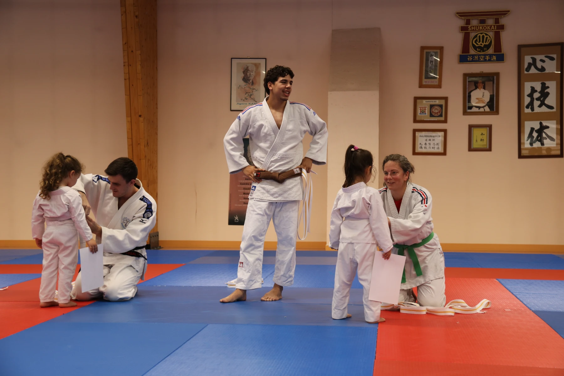 Un judoka ceinture marron souriant entouré d'enfants et d'une maman pendant la remise des ceintures.