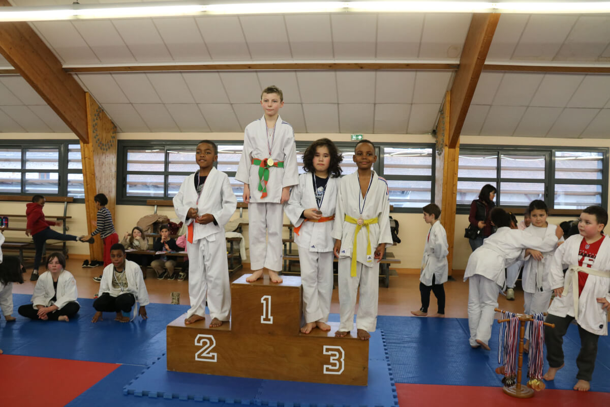 Démonstration de judo où un pratiquant ceinture noire exécute une projection sur un autre judoka, sous les regards d'enfants assis.
