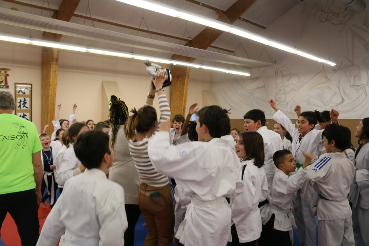Des enfants alignés en tenue de judo, attendant leur tour pour une activité, avec des ceintures colorées exposées devant eux.